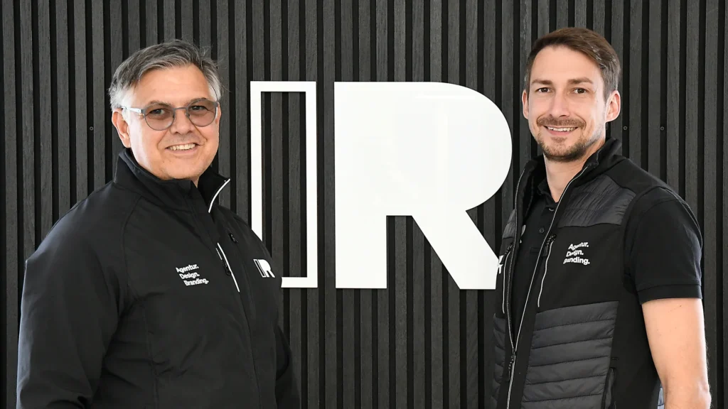 Zwei Personen stehen nebeneinander vor einer schwarzen Wand mit dem Logo „IR“.