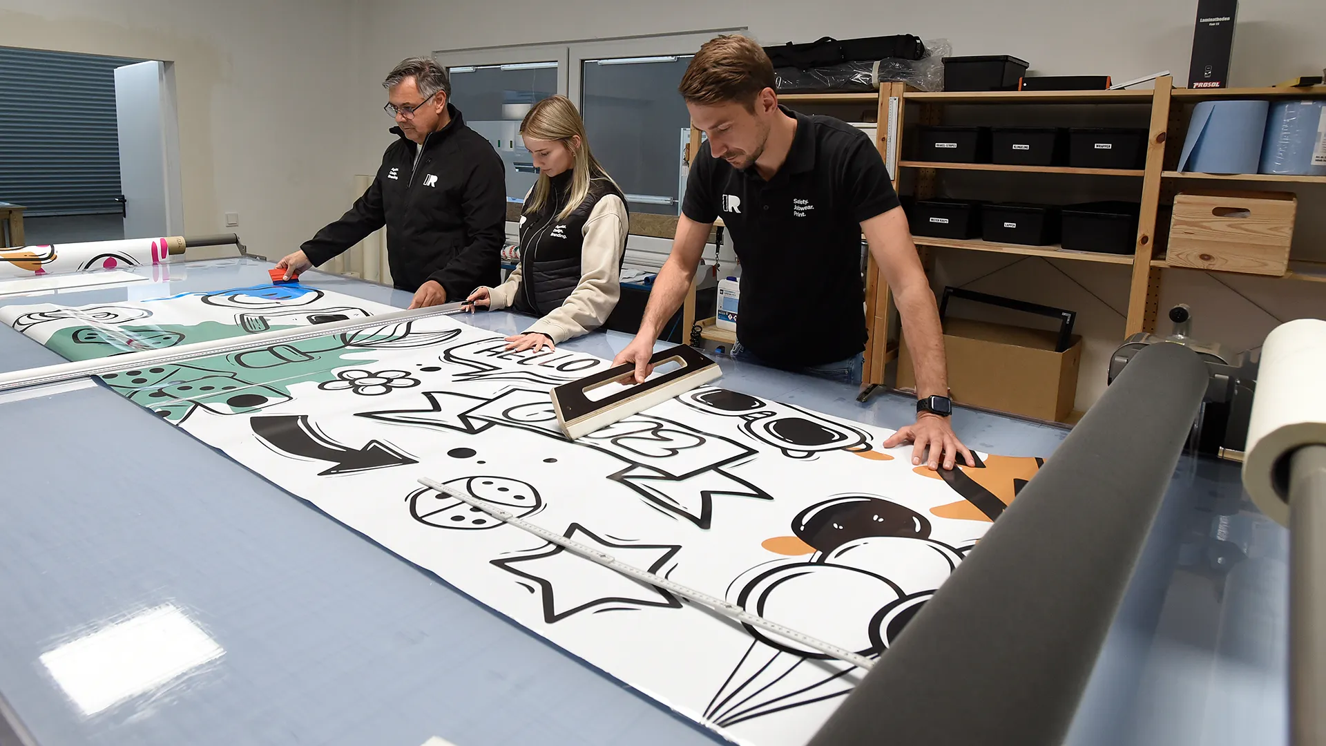 Drei Personen arbeiten gemeinsam an einem großen Tisch in einem modernen Arbeitsraum. Auf dem Tisch liegt eine großformatige, bunte Grafik mit Comic-Illustrationen, die von den Personen mit Werkzeugen geglättet und bearbeitet wird. Im Hintergrund stehen Regale mit Kisten, Papierrollen und weiteren Materialien. Die Szene vermittelt den Eindruck eines kreativen Arbeitsprozesses, etwa in einer Druckerei, Werbetechnik- oder Designwerkstatt.